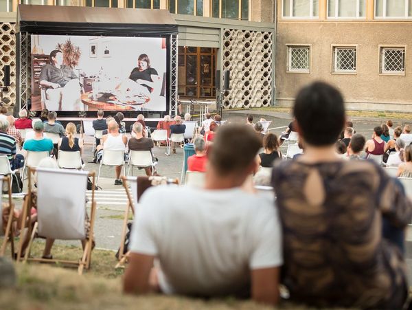 Zuschauer sitzen auf Stühlen und auf der Wiese und schauen in Richtung einer großen Leinwand.