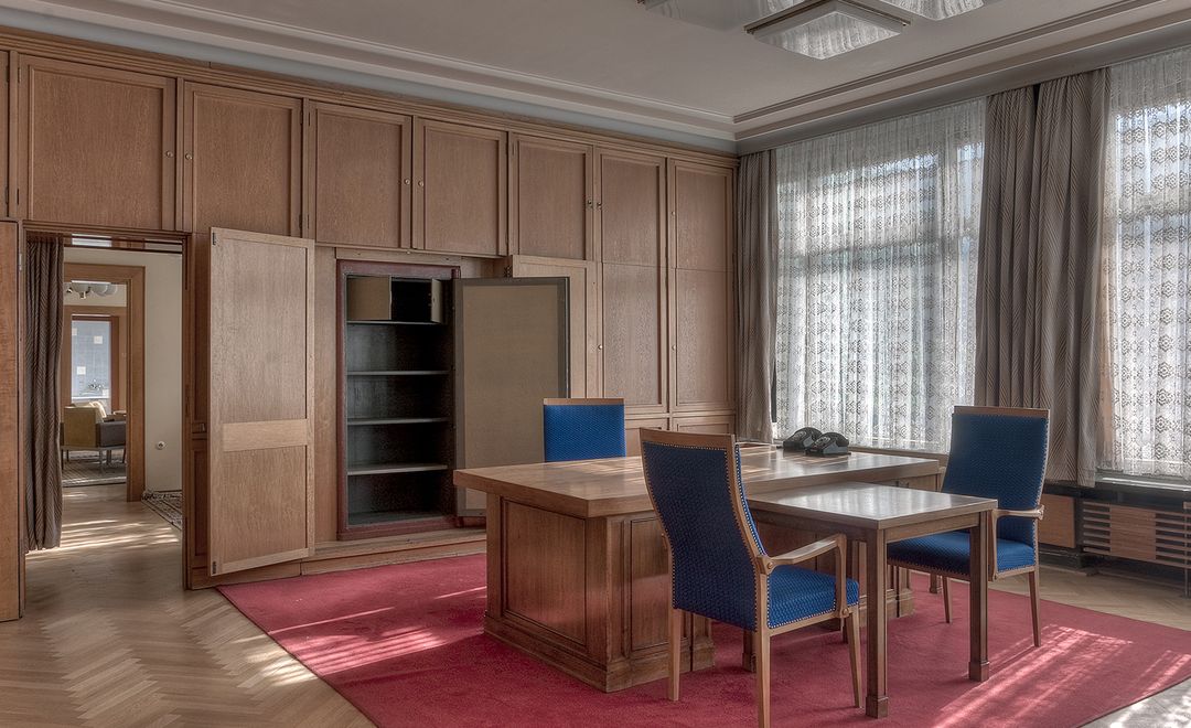 Das Bild zeigt ein hochwertig eingerichtetes Büro mit Holzverkleidung. In der Mitte des Raumes steht ein großer Schreibtsich mit drei Stühlen. Die Fenster sind mit halbdurchlässigen Vorhängen abgedeckt