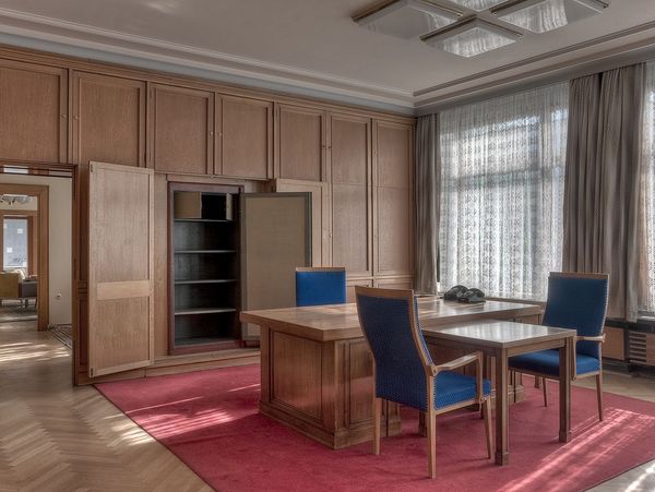 Das Bild zeigt ein hochwertig eingerichtetes Büro mit Holzverkleidung. In der Mitte des Raumes steht ein großer Schreibtsich mit drei Stühlen. Die Fenster sind mit halbdurchlässigen Vorhängen abgedeckt